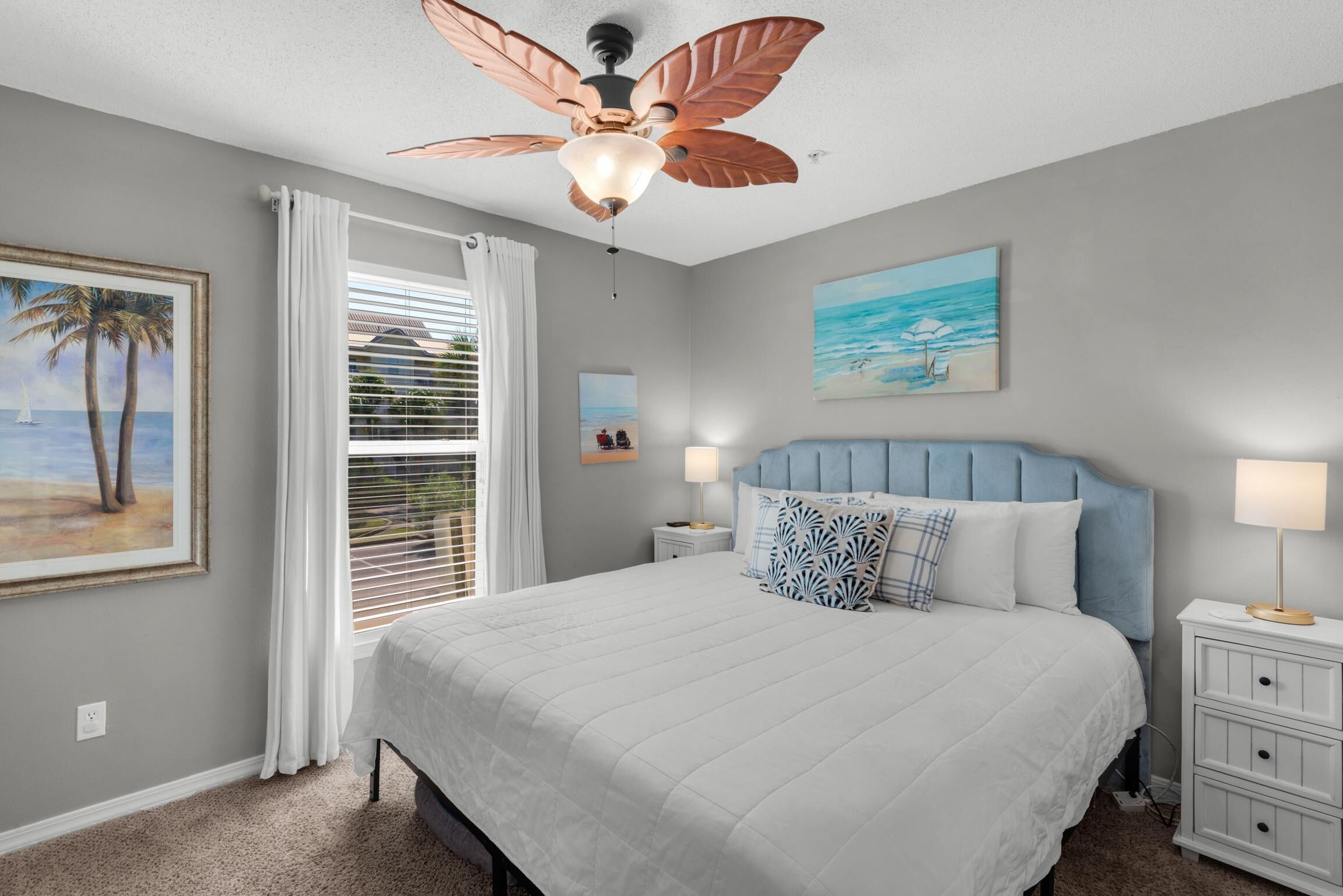 A bedroom with a bed, a ceiling fan, a nightstand, a lamp, and a painting of a beach scene.