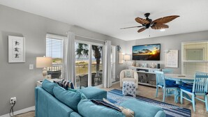 A modern living room with a blue sofa, a ceiling fan, a dining area with blue chairs, and a view of the beach through large windows.