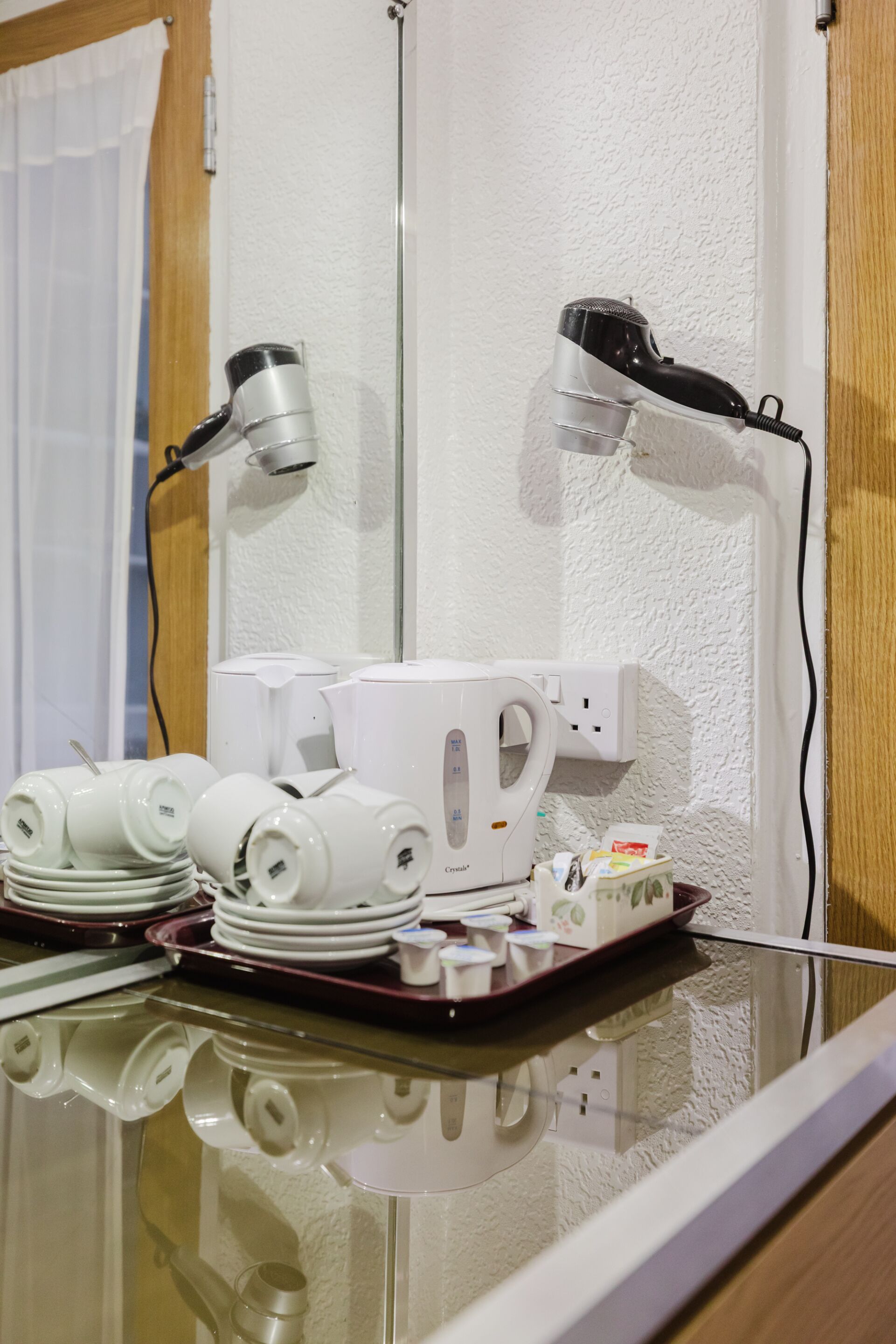 A hotel room with a glass table displaying a hairdryer, a kettle, and a tray with plates and cups.