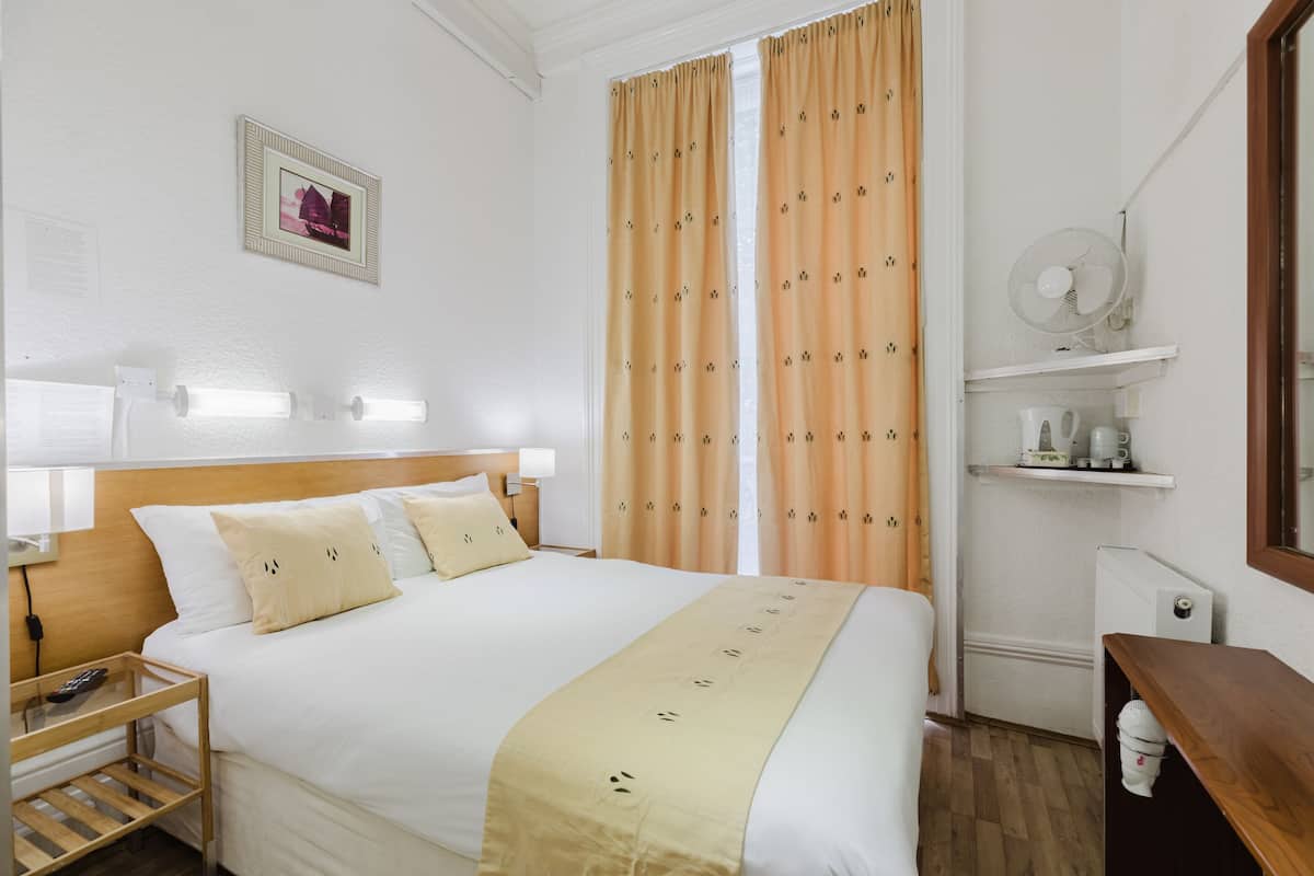 A neatly arranged hotel room with a large bed, a bedside table with a lamp, a small shelf with toiletries, and a window with curtains.
