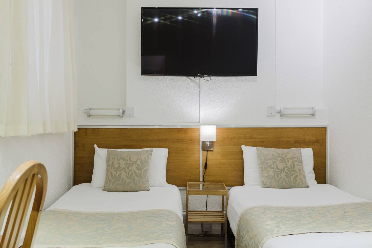 A hotel room with two beds, a TV mounted on the wall, and a nightstand with a lamp.