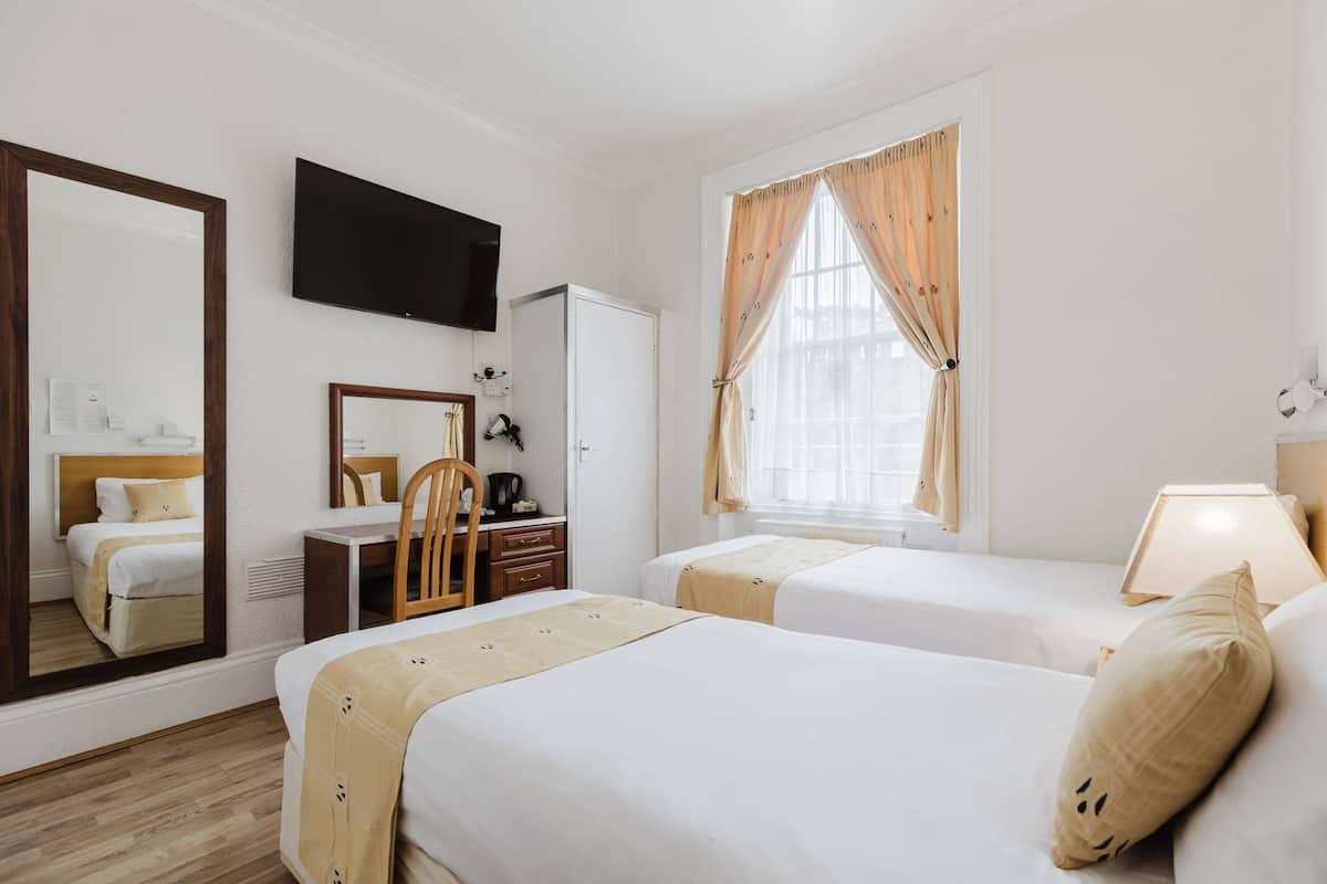 A hotel room with two beds, a TV mounted on the wall, a desk with a chair, and a window with curtains.