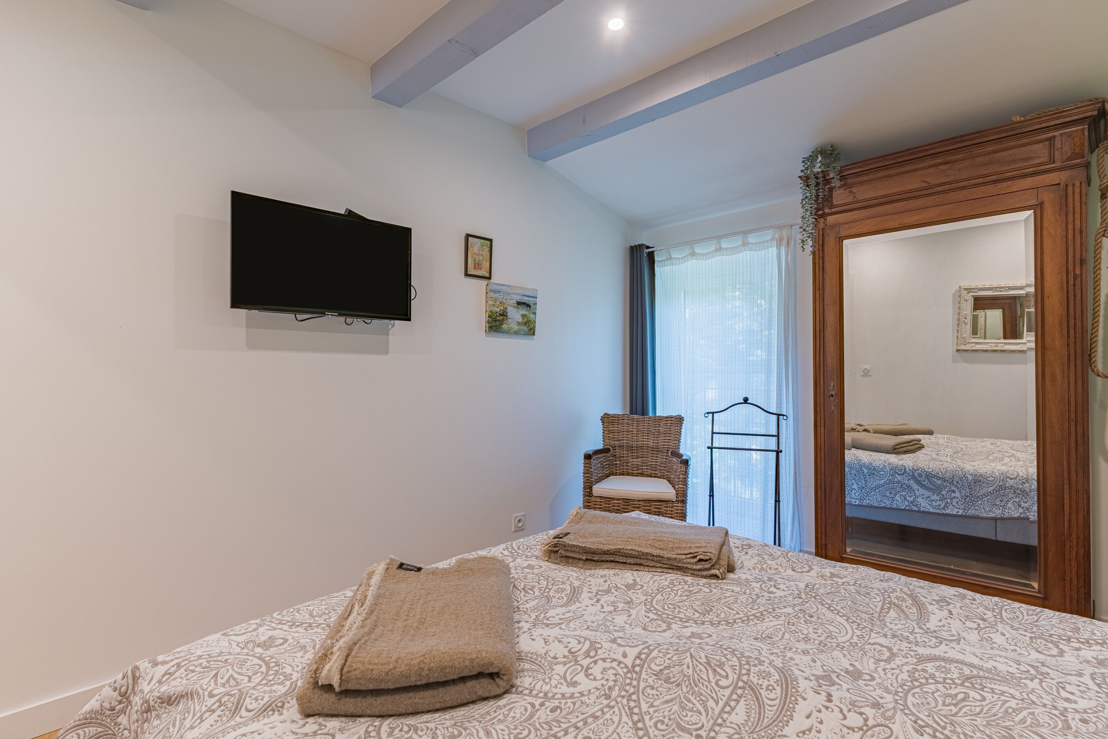 A bedroom with a bed, a television, a chair, a mirror, and a window with curtains.