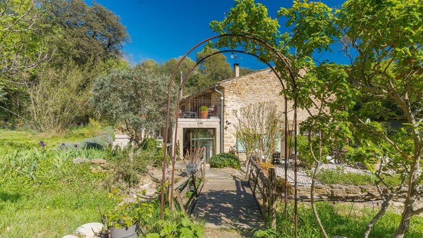 A stone house with a wooden deck, surrounded by lush greenery and a clear blue sky.
