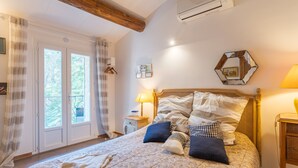 Une chambre avec un grand lit, une poutre en bois au plafond, une fenêtre avec des rideaux, une table de chevet avec une lampe et un climatiseur fixé au mur.