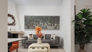 A living room with a grey sofa, a white coffee table with orange flowers, a large abstract painting, and a potted plant. - Hera Family Apartment, Olympian Zeus Apartments (Athens)