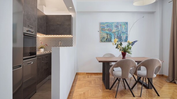 A modern kitchen with dark cabinets, a built-in oven, and a herringbone-patterned backsplash. There is a dining area with a wooden table and four chairs, and a painting on the wall. - Zeus Grand Apartment, Olympian Zeus Apartments (Athens)