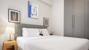 A neatly made bed with white bedding, a grey headboard, and a bedside table with a lamp. There are two framed pictures on the wall above the bed. - Zeus Grand Apartment, Olympian Zeus Apartments (Athens)