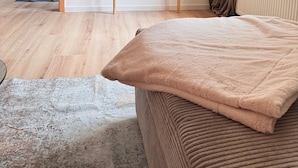 Une chambre moderne avec un grand téléviseur à écran plat fixé sur une console blanche, un lit recouvert d’une couverture beige et une vue sur un paysage verdoyant par la fenêtre.