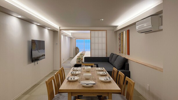 A dining area with a long table set for four, wooden chairs, a sofa, and a TV on the wall. - bHotel Miyajima Omotesando 21 (Hatsukaichi)