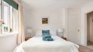 A neatly arranged bedroom with a bed, bedside tables, a window with curtains, a door, and a wall-mounted picture. - The Waterside Apartment, ideal for families (Largs)