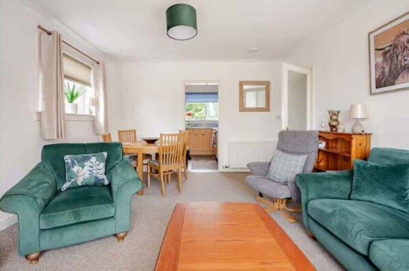 A living room with two green sofas, a wooden coffee table, a dining table with chairs, and a kitchen visible through an open doorway. - The Waterside Apartment, ideal for families (Largs)