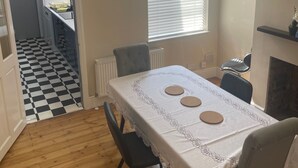 Un coin repas avec une table dressée pour quatre personnes, une cheminée et une cuisine visible à travers une porte ouverte.