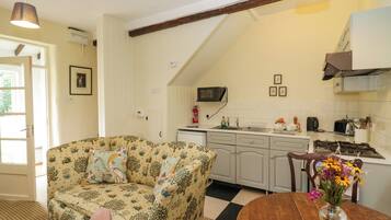 A cozy living room with a floral sofa, a dining table with a vase of flowers, and a kitchen with white cabinets and a microwave.