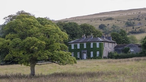 Una casa grande con fachada verde, rodeada por un campo cubierto de hierba y una colina al fondo.