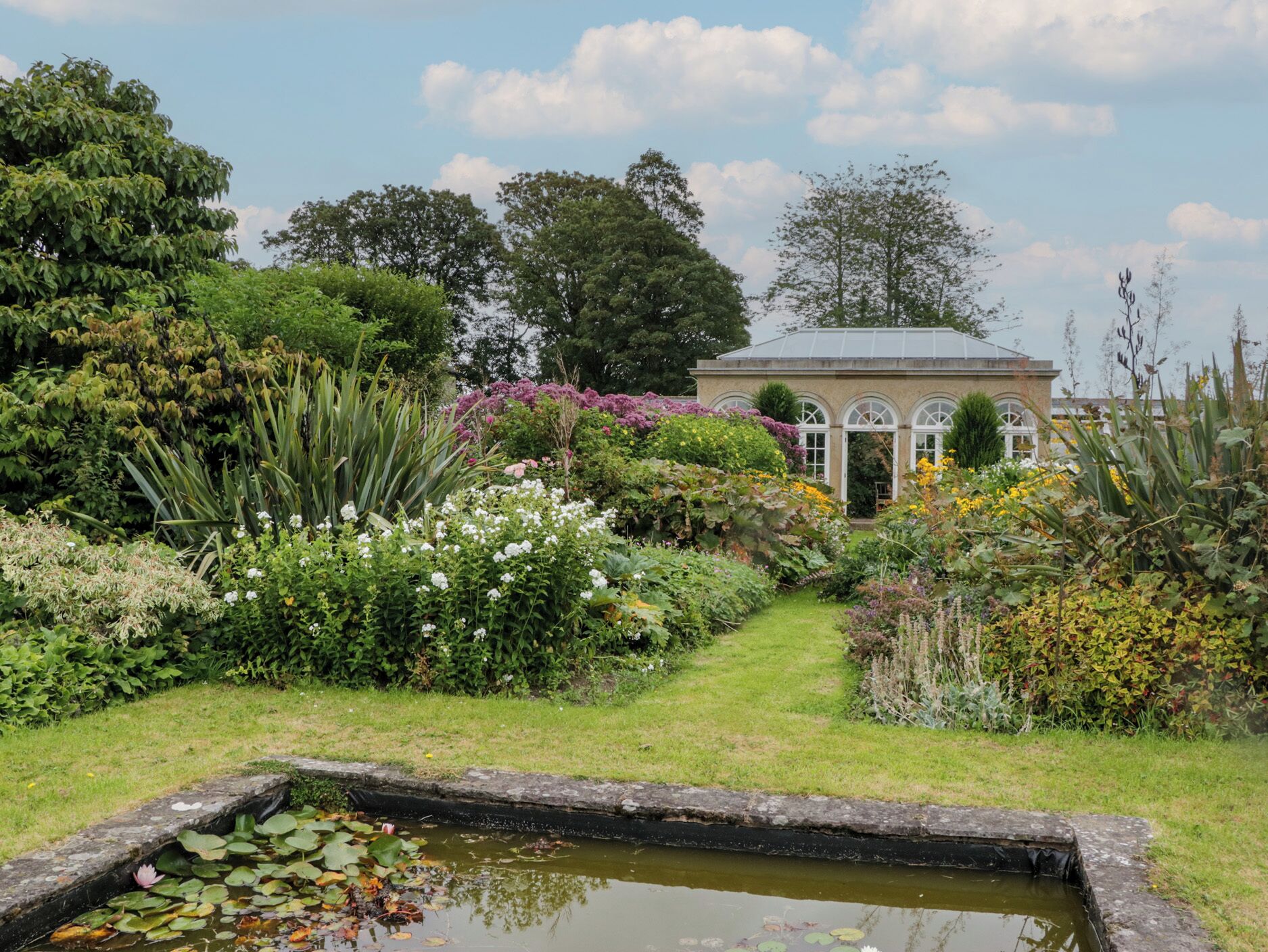 Un jardín con un estanque, diversas plantas y un invernadero.
