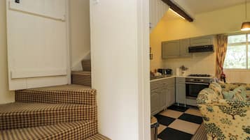Cozinha com fogão a gás, forno e janela com cortinas. Degraus levam a uma porta com moldura branca.