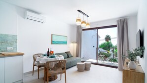 Uma sala de estar moderna com sofá, mesa de jantar e cadeiras. Há uma TV na parede, uma janela com cortinas e uma varanda com mobiliário de exterior.