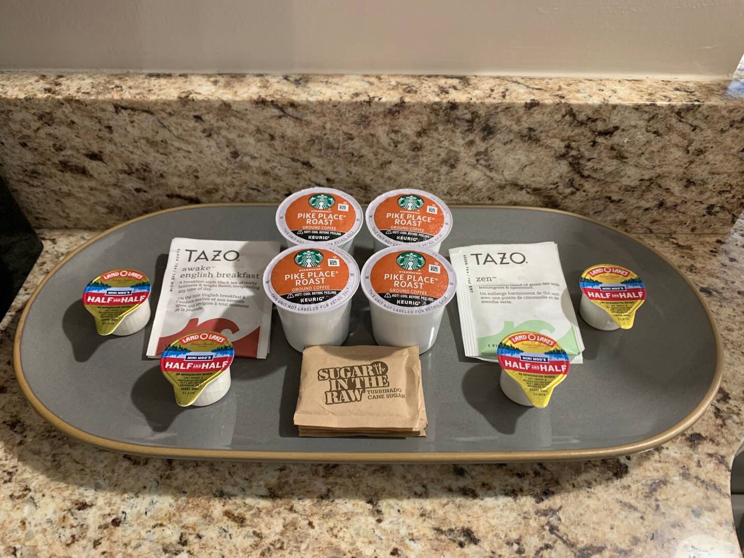 A tray with Starbucks Pike Place Roast coffee cups, Tazo tea bags, and Half & Half dairy products on a granite countertop.