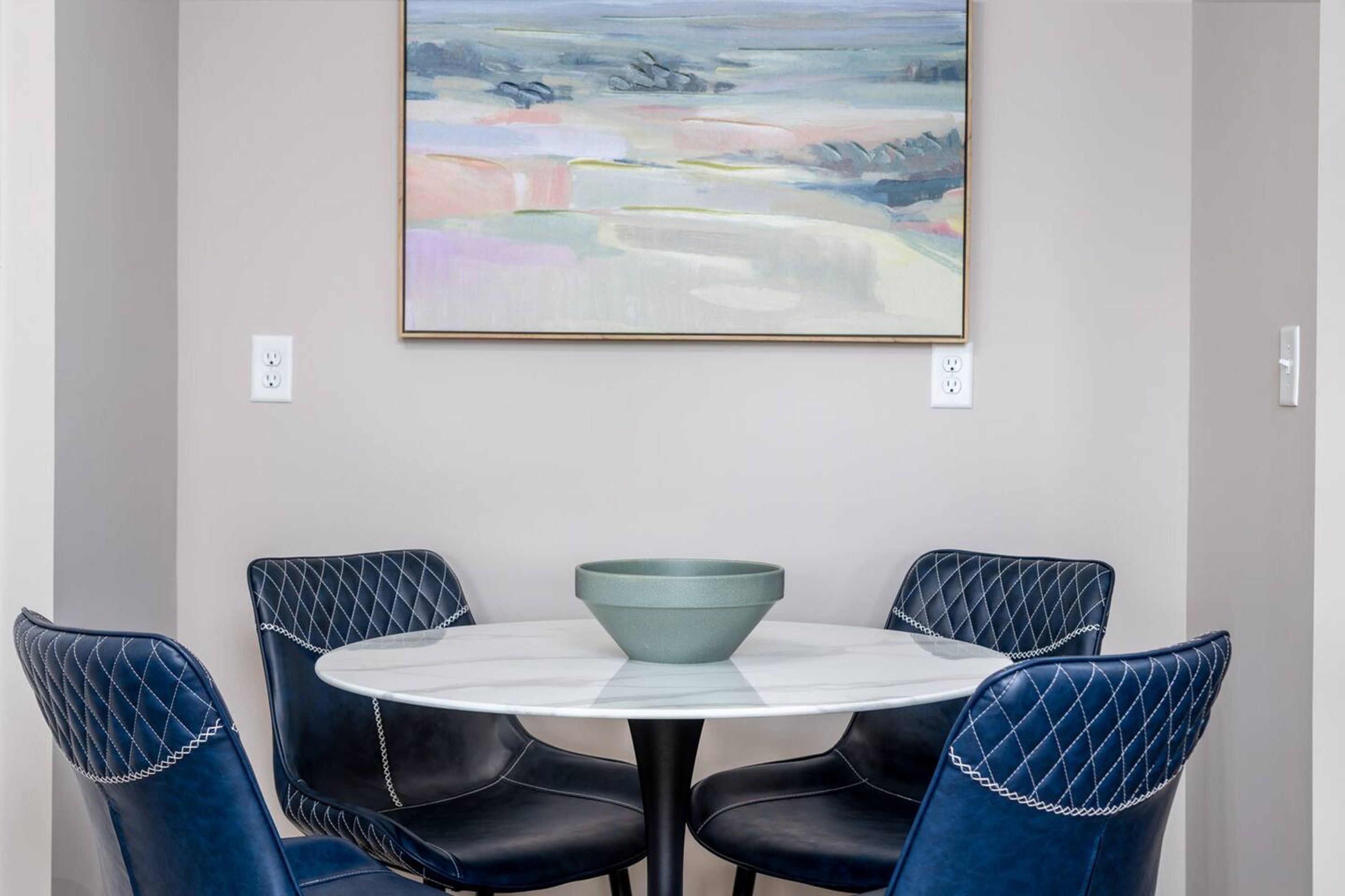 A dining area with a round table and four chairs, a bowl on the table, and a painting on the wall.