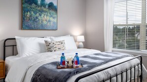 A neatly made bed with white bedding, a painting above it, and a window with blinds. - Top Floor Water View King Bed Pool Gym (Westerville)
