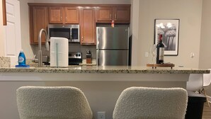 A kitchen with wooden cabinets, stainless steel appliances, a bar area with two plush chairs, and a wine bottle on the counter. - Polaris King Flat Pet-friendly Nature Views (Westerville)