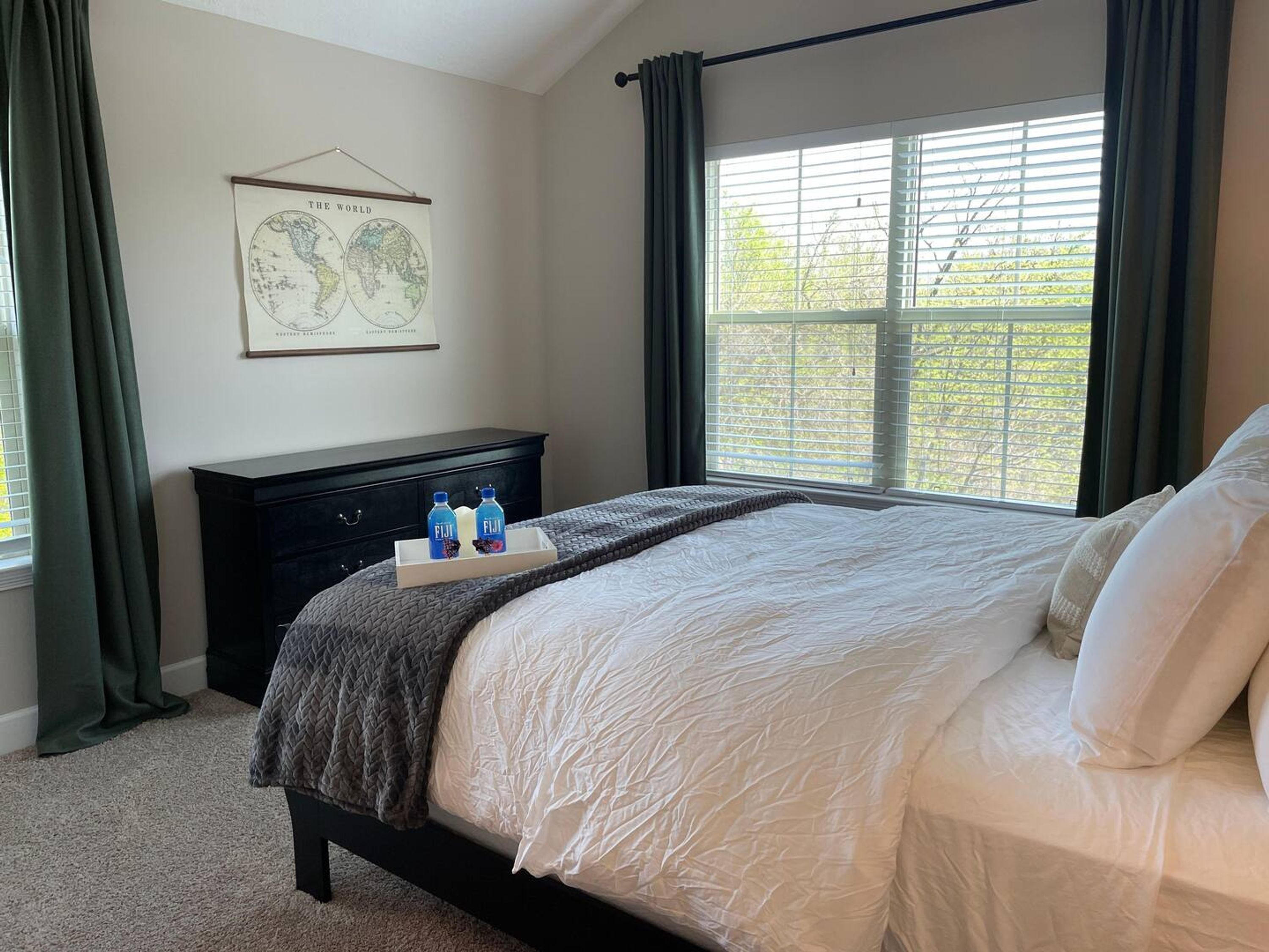 A bedroom with a bed, a dresser, a map on the wall, and a window with curtains.