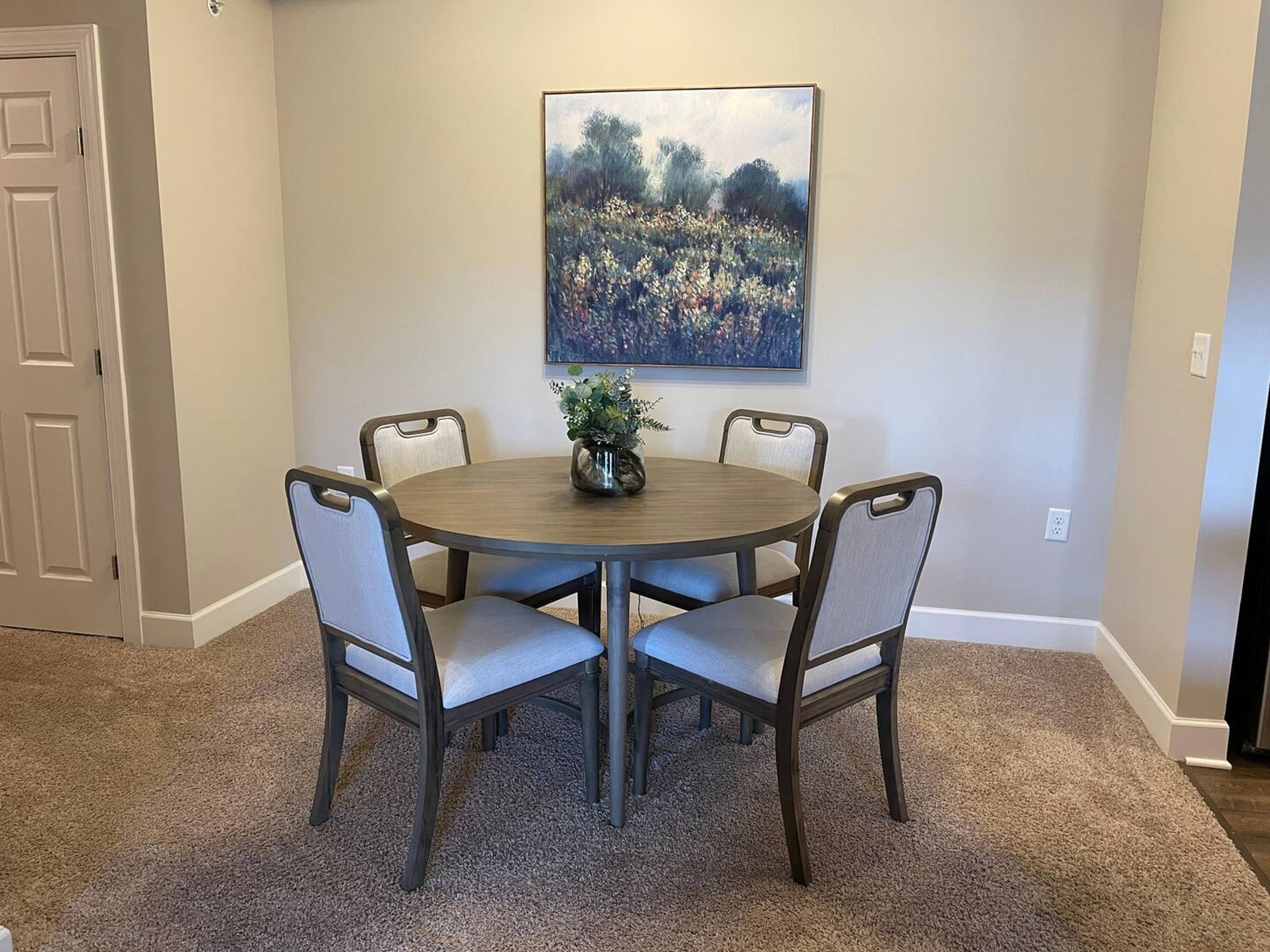 A dining area with a round table, four chairs, and a painting on the wall.