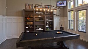 A pool table with a hanging light fixture, surrounded by shelves with various items.