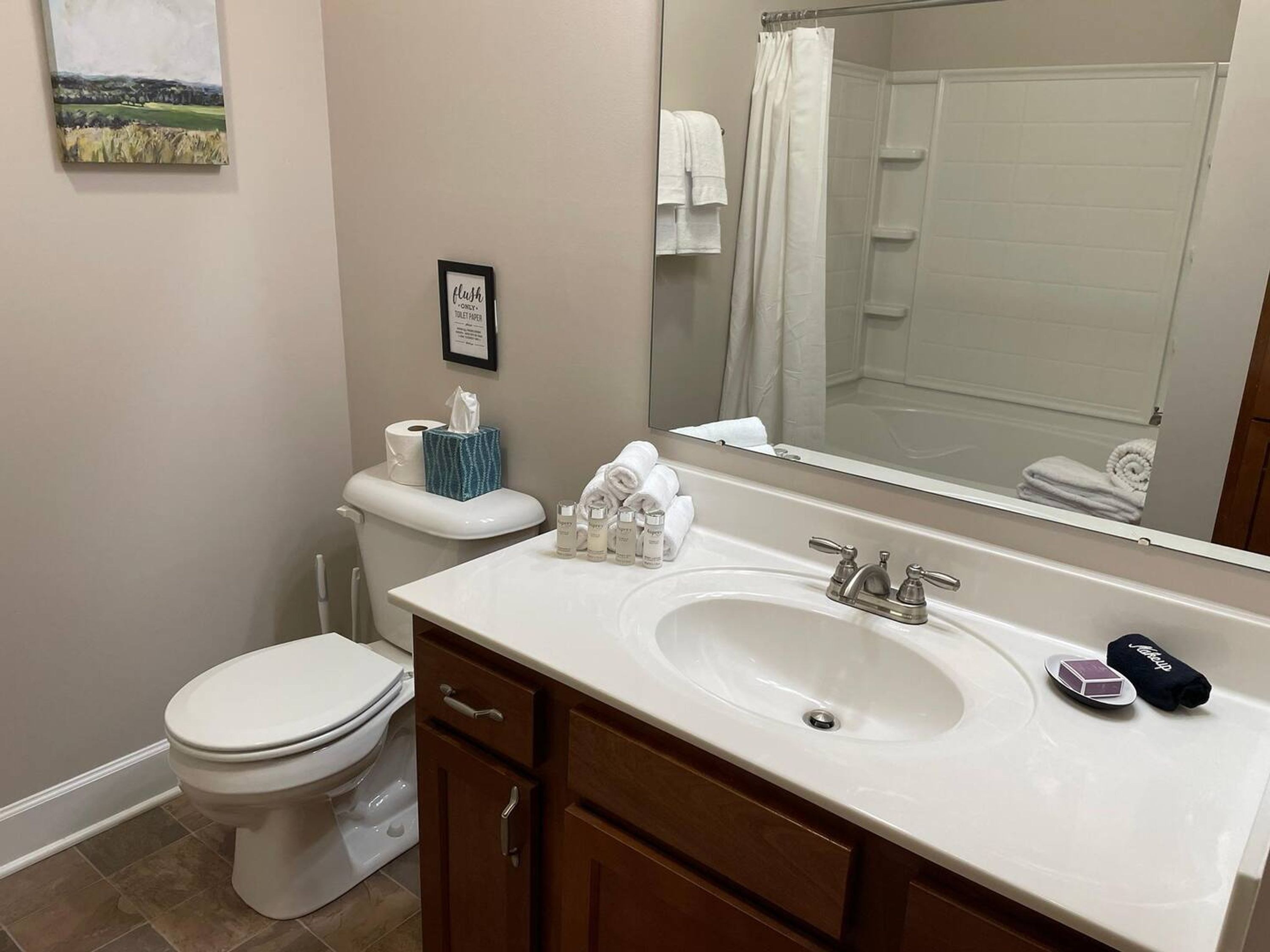 A bathroom with a toilet, sink, and shower. There is a framed sign on the wall, a towel rack, and a mirror reflecting the shower area.