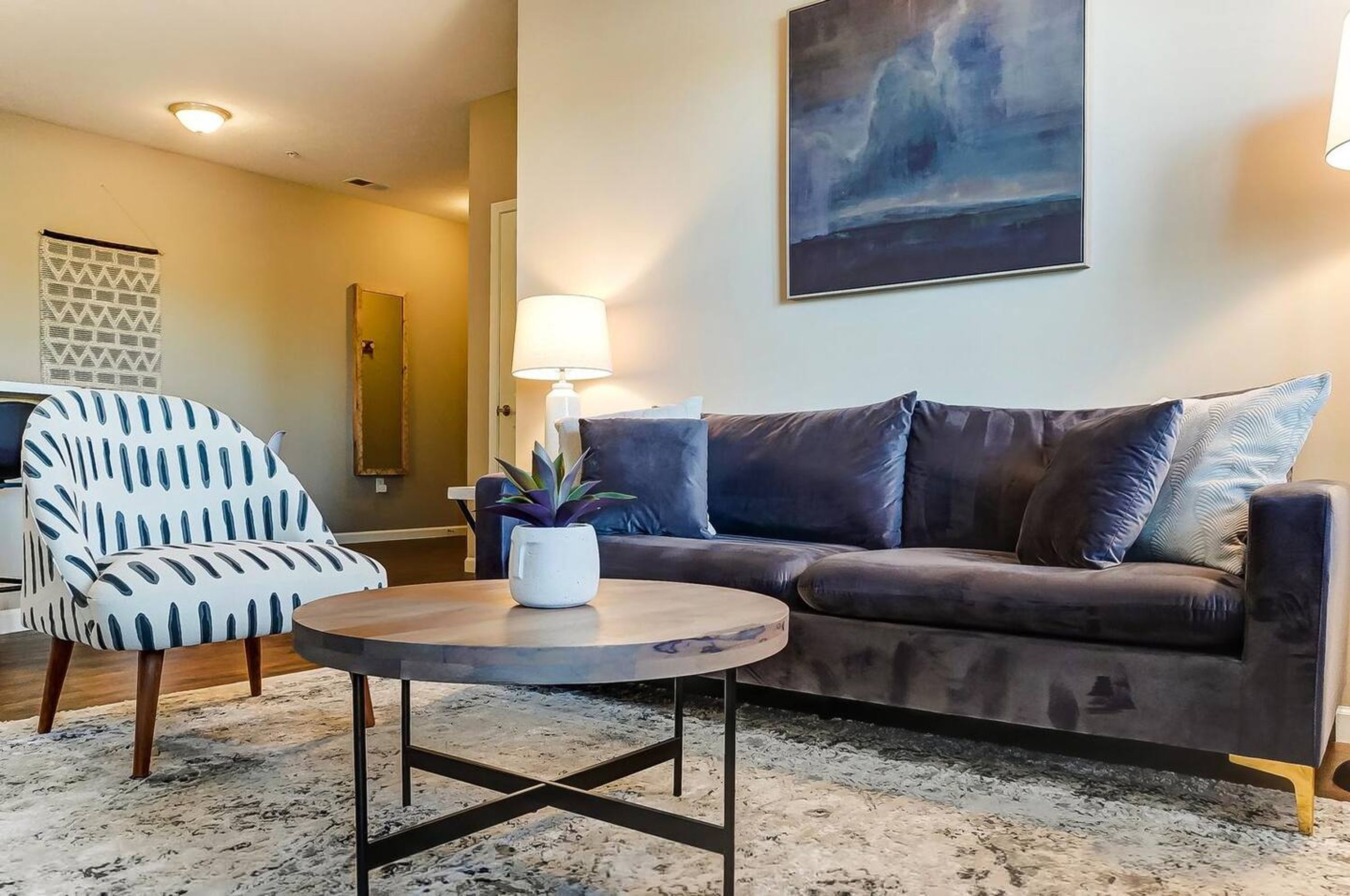 A living room with a brown leather sofa, a patterned armchair, a round coffee table with a plant, and a painting on the wall.
