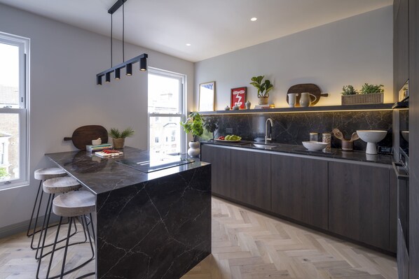 A modern kitchen with a dark marble island, bar stools, and a herringbone wood floor.