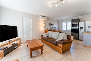 Una sala de estar con televisor, mesa de centro de madera, sofá de cuero marrón y una zona de cocina con refrigerador y estufa.