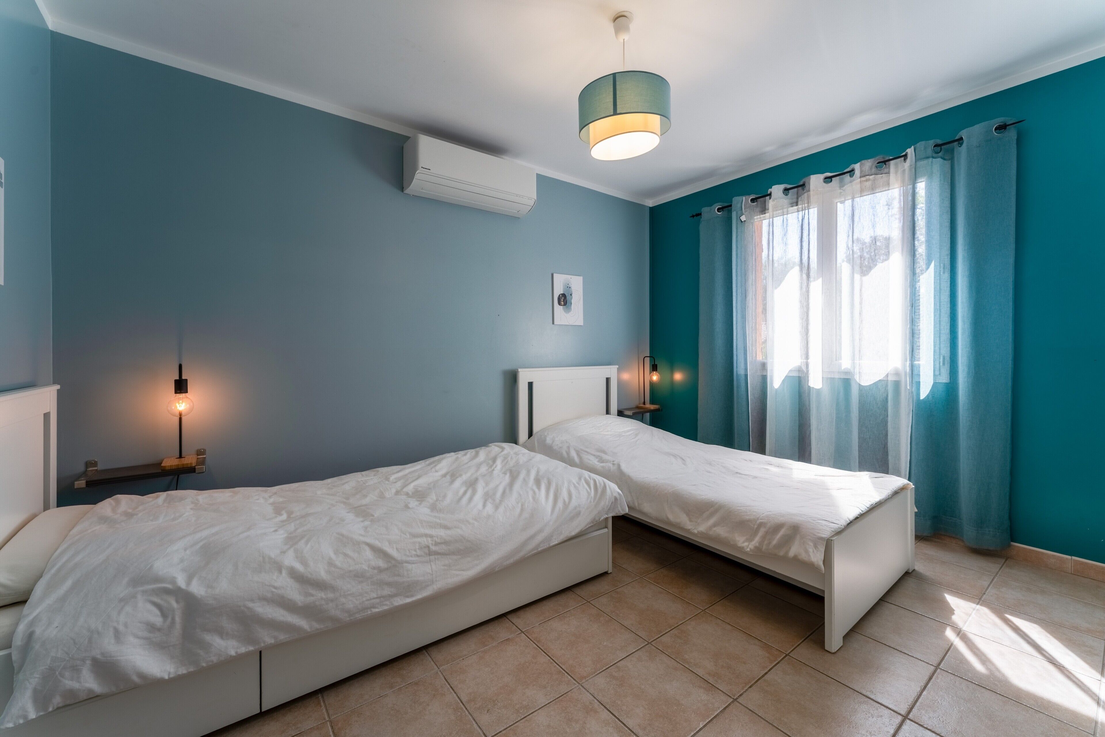 A bedroom with two beds, a ceiling light, an air conditioner, and a window with curtains.