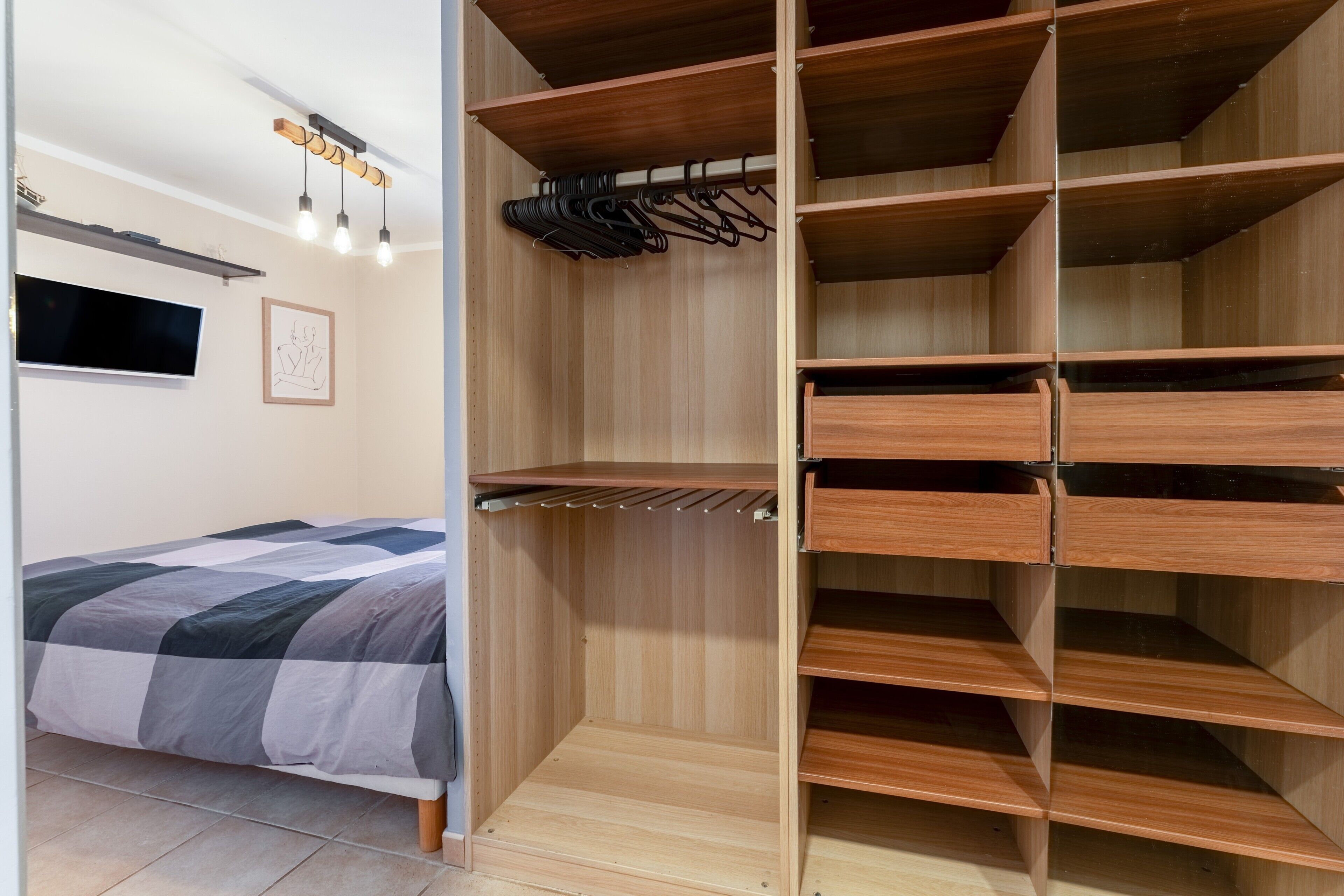 Un dormitorio con una cama, un armario de madera con estantes y espacio para colgar, un televisor montado en la pared y un cuadro enmarcado.