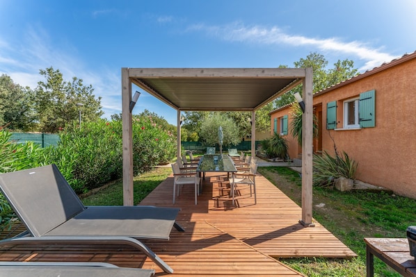 Una terraza de madera con una mesa y sillas de vidrio, rodeada de follaje y un edificio con contraventanas verdes.