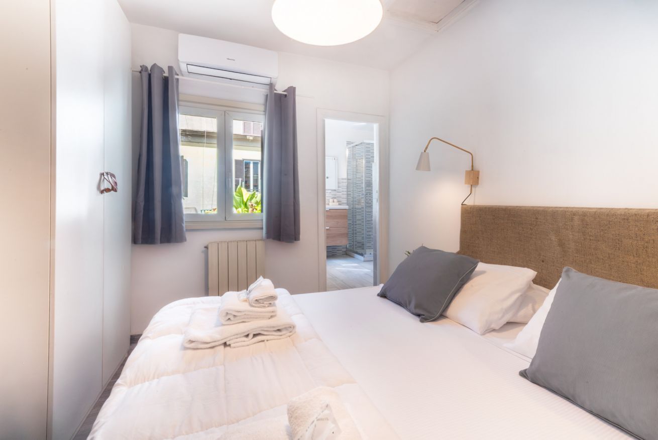 A hotel room with a bed, a window with curtains, an air conditioning unit, and a wall-mounted lamp.