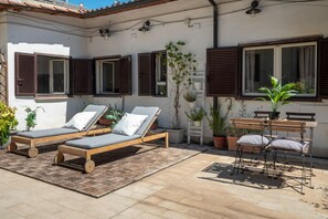 A patio with two wooden sun loungers, a wooden table with chairs, and potted plants.