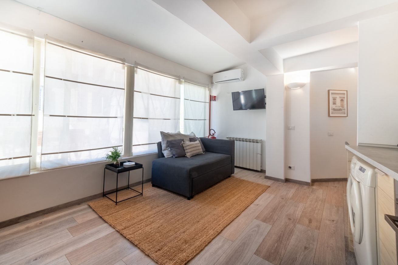 A modern living room with a sofa, a small side table, a large window, a TV, and a washing machine.