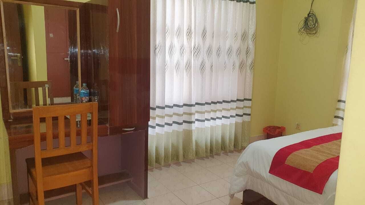 A room with a bed, a wooden chair, a desk, a mirror, and a window with patterned curtains.