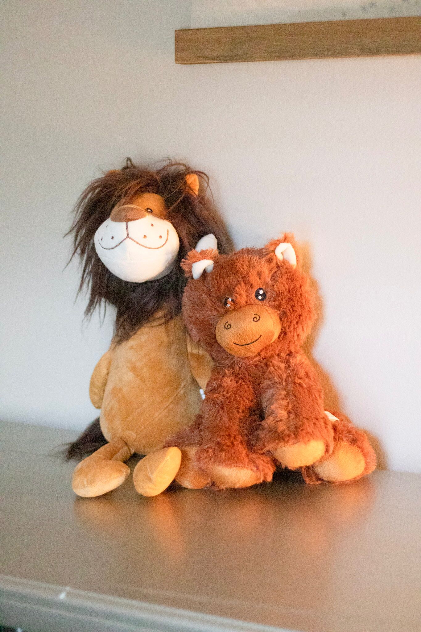 Two plush toys, one resembling a lion and the other a bear, sitting on a shelf against a plain wall.