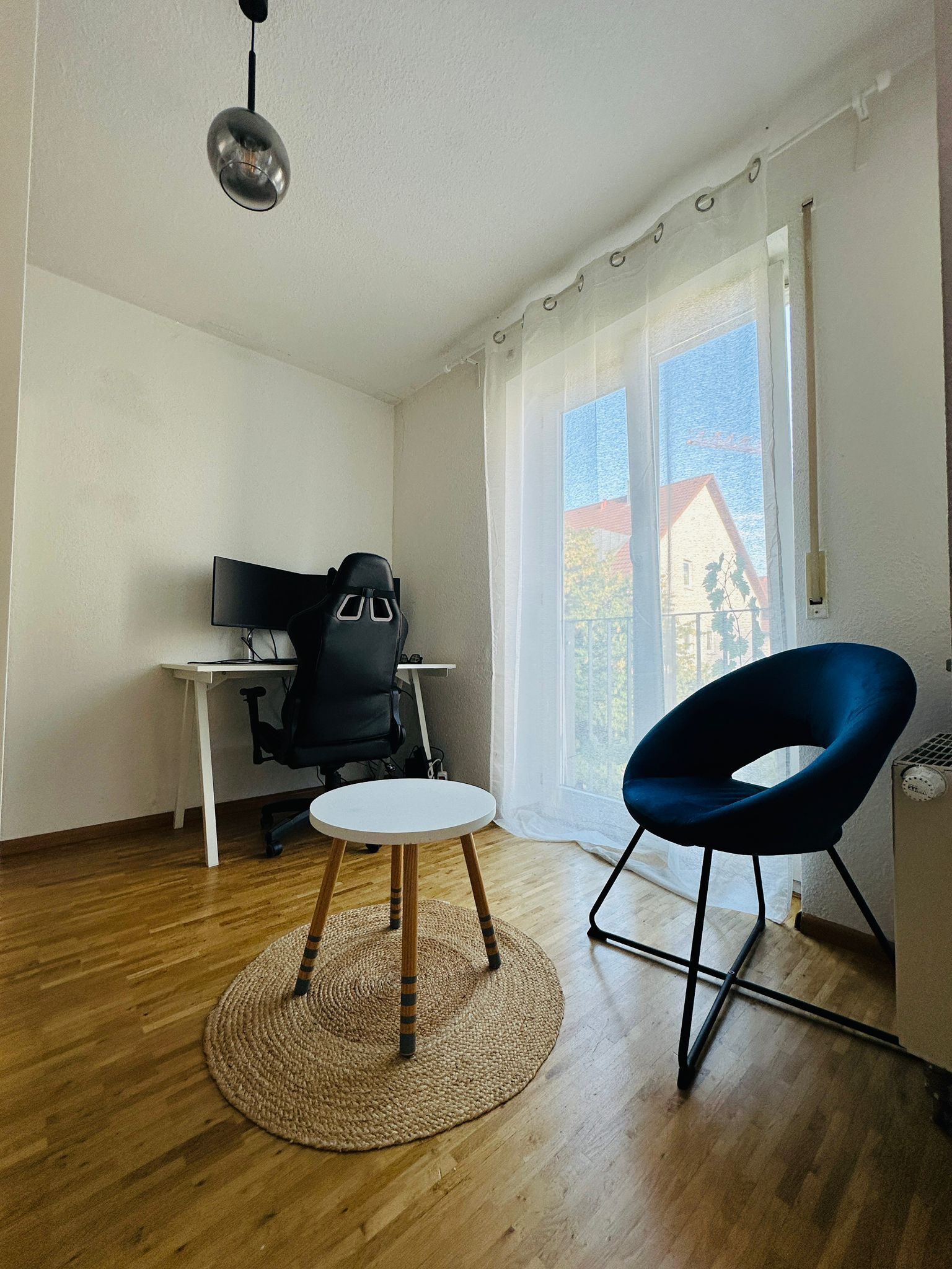 Une pièce avec un plancher en bois, un tapis rond tissé, un fauteuil de bureau noir, une table blanche, une chaise bleue, un luminaire suspendu et une fenêtre avec des rideaux.