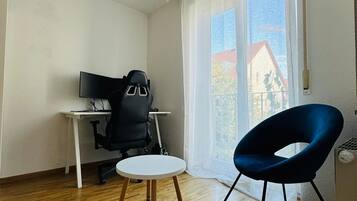 Une pièce avec un plancher en bois, un tapis rond tissé, un fauteuil de bureau noir, une table blanche, une chaise bleue, un luminaire suspendu et une fenêtre avec des rideaux.