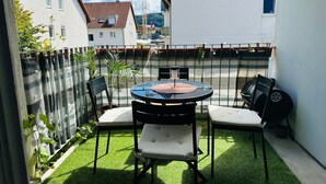 Een klein terras buiten met een ronde tafel en vier stoelen, omringd door een lage haag en een gazon.