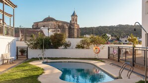 Une piscine entourée d’une terrasse, avec un banc et une vue sur un bâtiment historique doté d’un clocher.