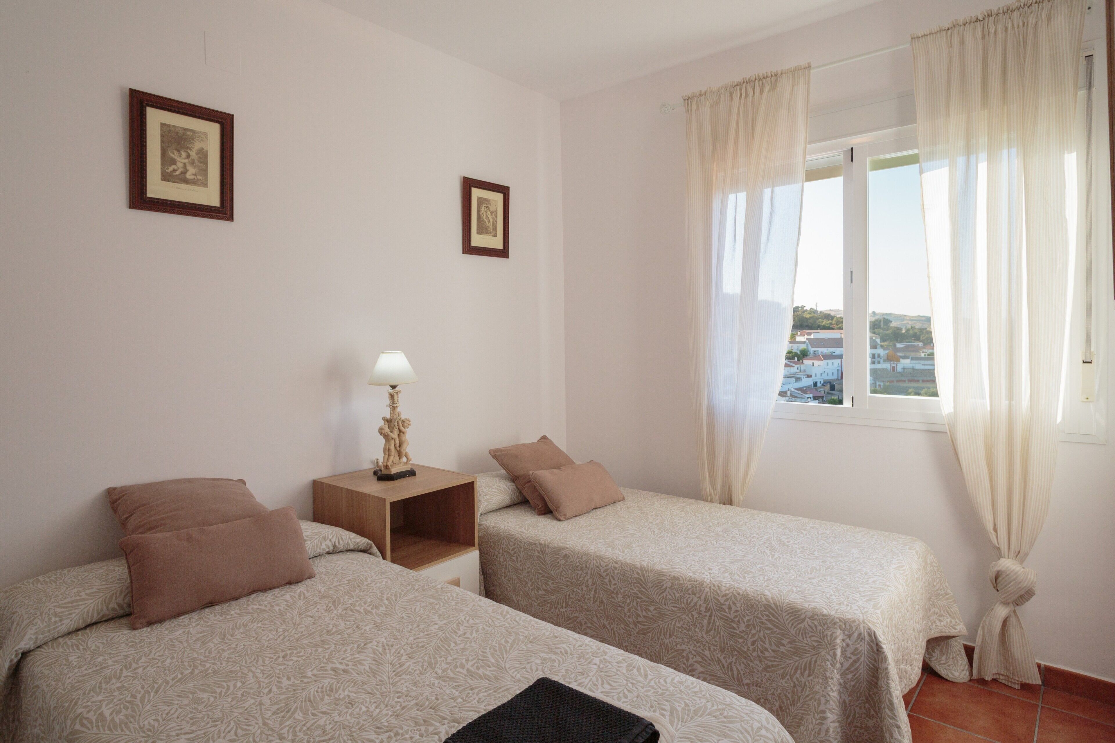 Quarto com duas camas, uma mesa de cabeceira com abajur e uma janela com cortinas.