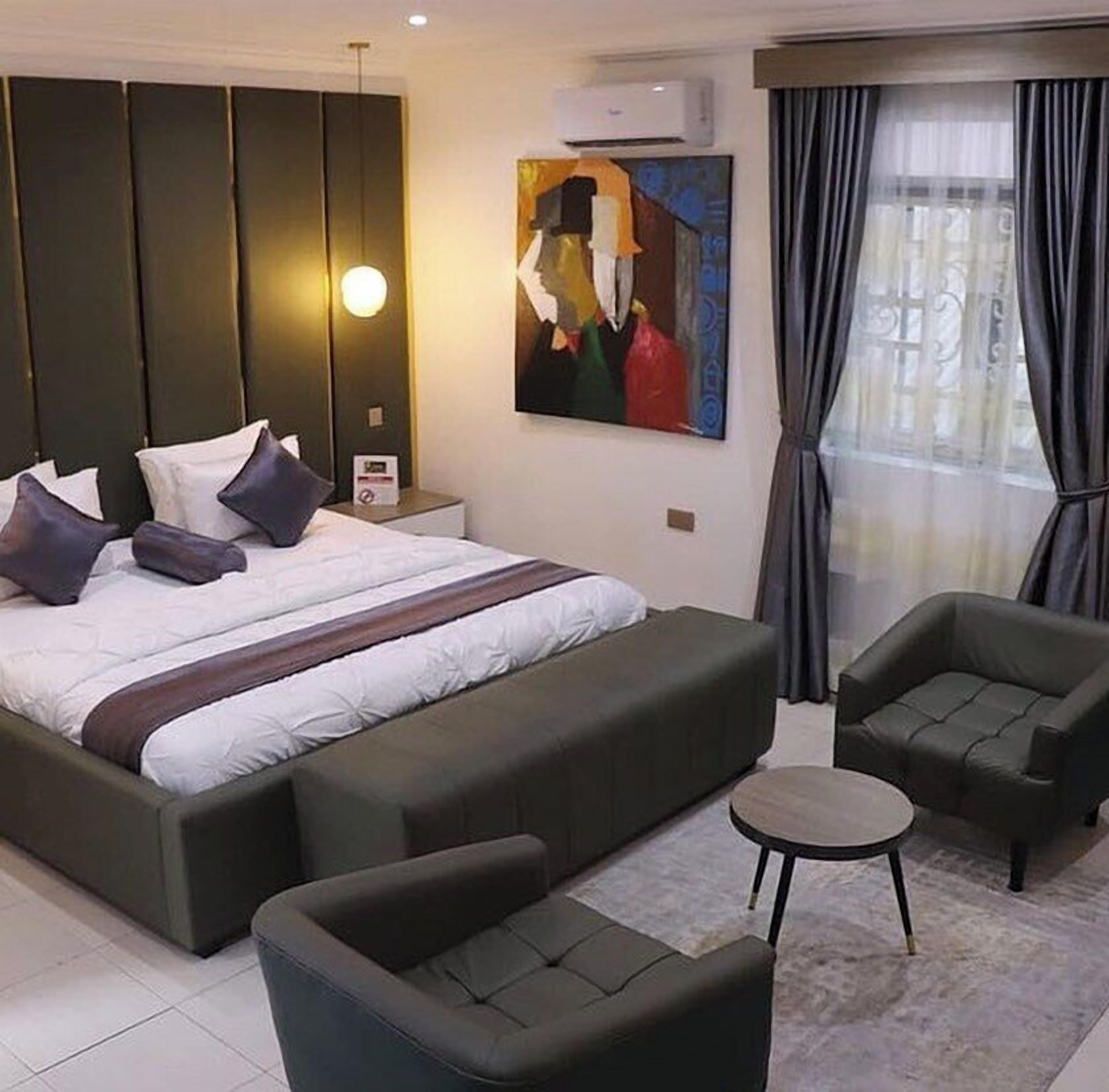 A modern hotel room with a large bed, a side table, a painting, a sofa, and a small round table.