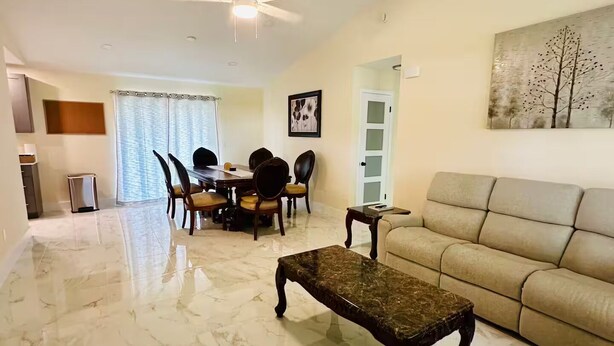 A spacious living room with a marble floor, a beige sofa, a dining area with a table and chairs, and a ceiling fan. - The 98 House Minutes from WEC, OBS, FAST & HITS. Located off of W HWY 40! (Ocala)