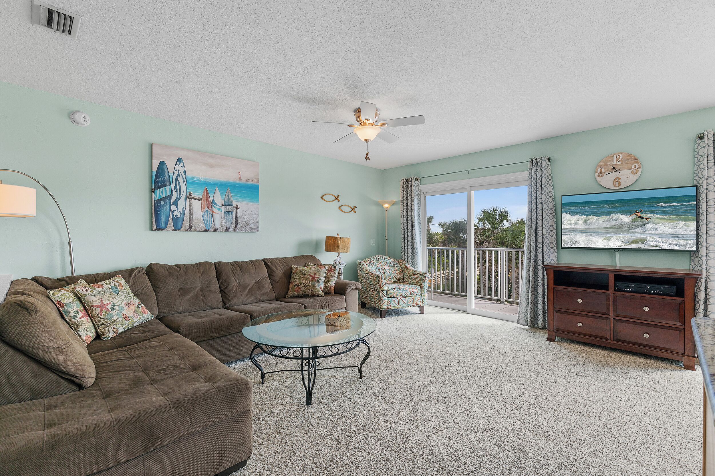 A living room with a brown sectional sofa, a glass coffee table, a TV on a wooden stand, a ceiling fan, and a painting of surfboards.