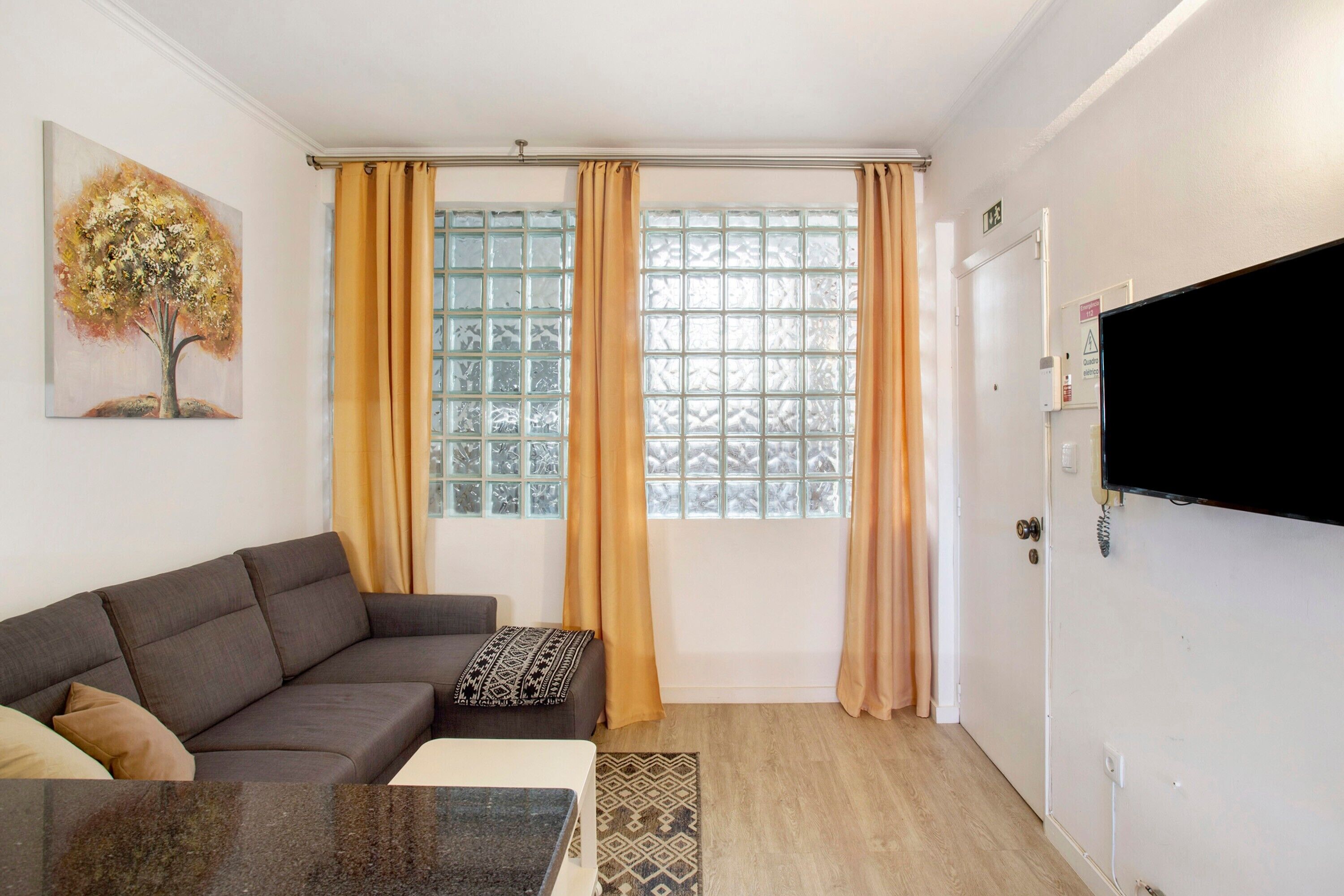 A living room with a sofa, a coffee table, a TV mounted on the wall, and a window with curtains.